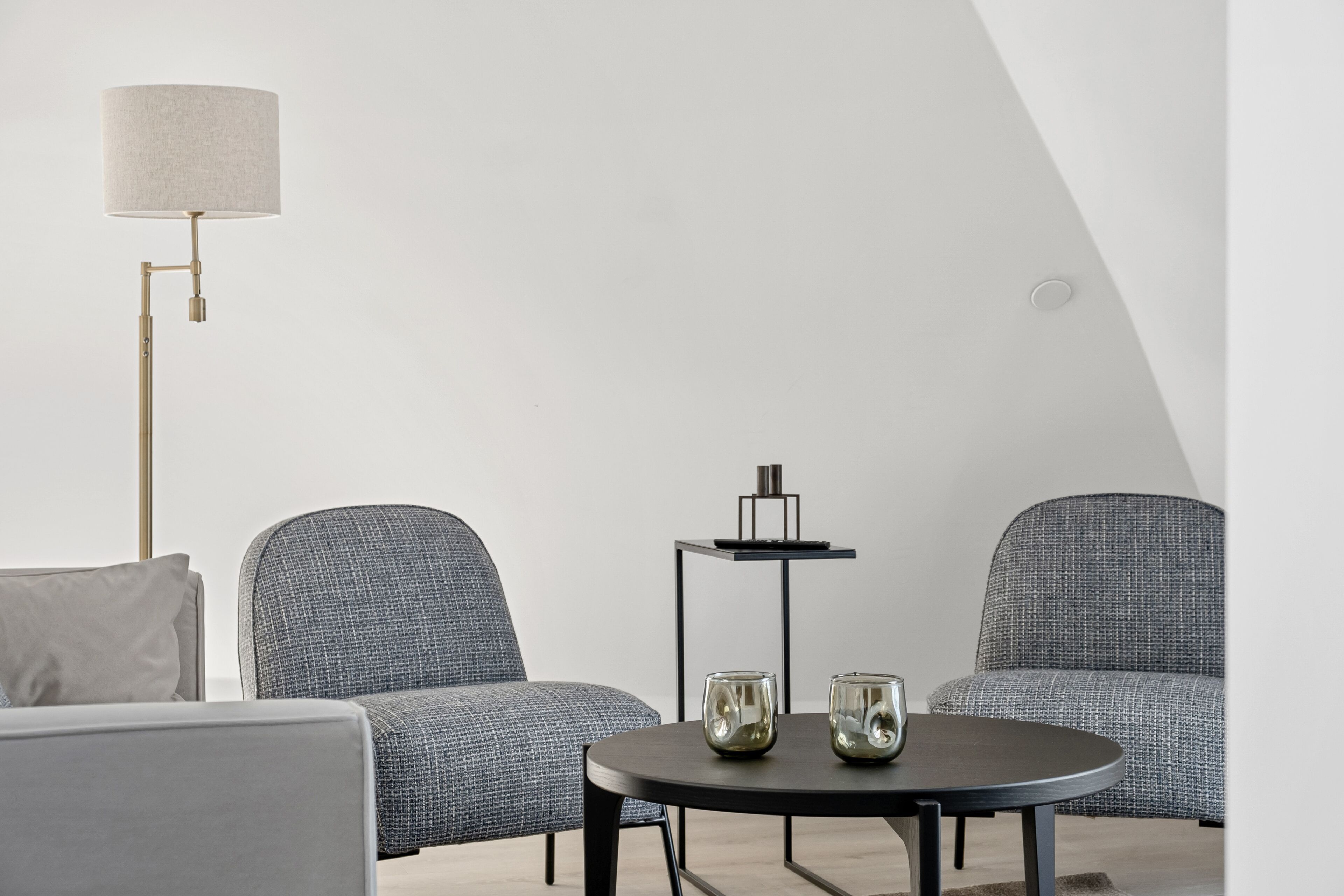 Ein modernes Zimmer mit zwei grauen Polsterstühlen, einem kleinen runden Tisch mit zwei Glasvasen und einer Stehlampe mit zylindrischem Lampenschirm.
