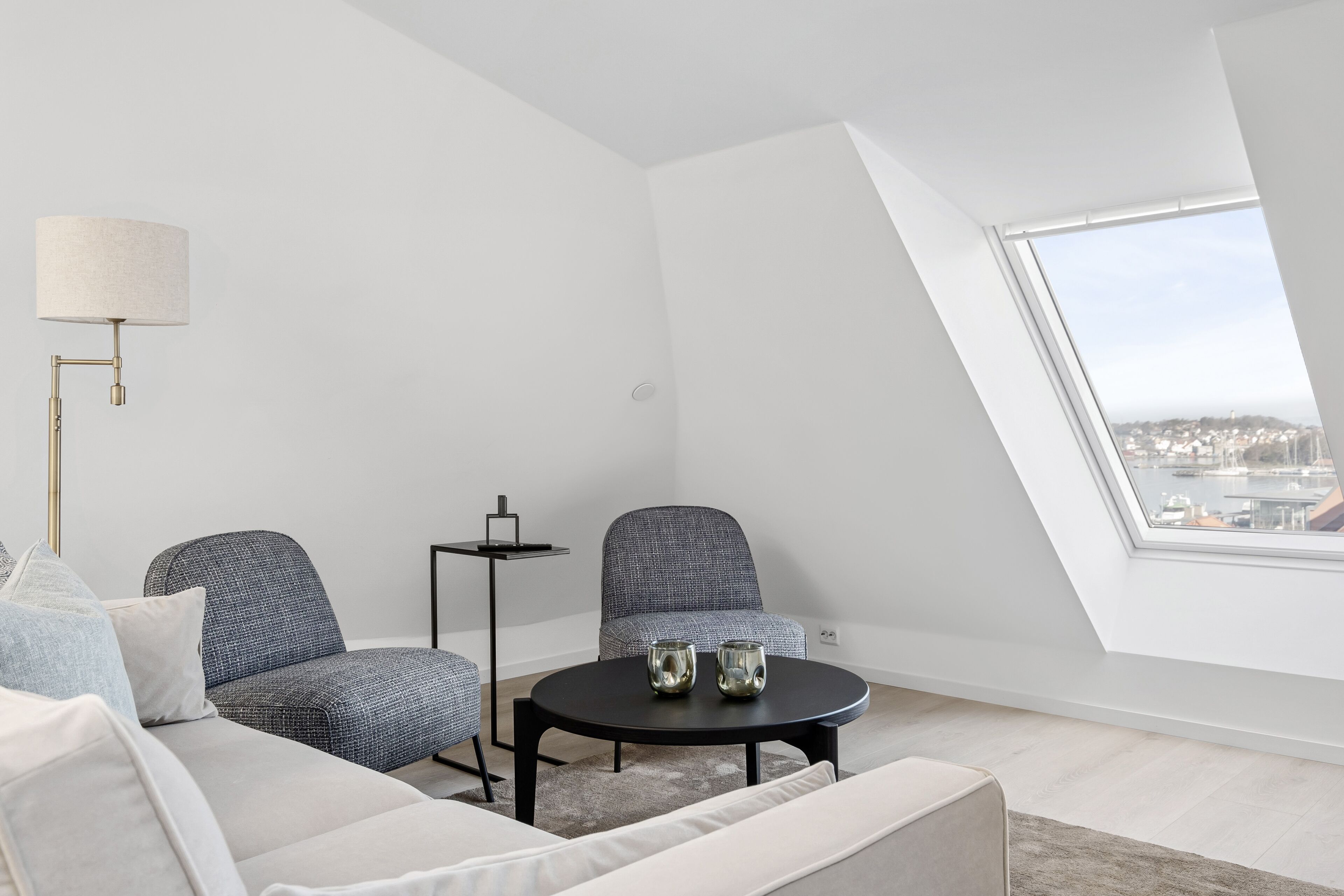 A modern living room with a high ceiling, a round black table, two grey armchairs, a white sofa, and a floor lamp.
