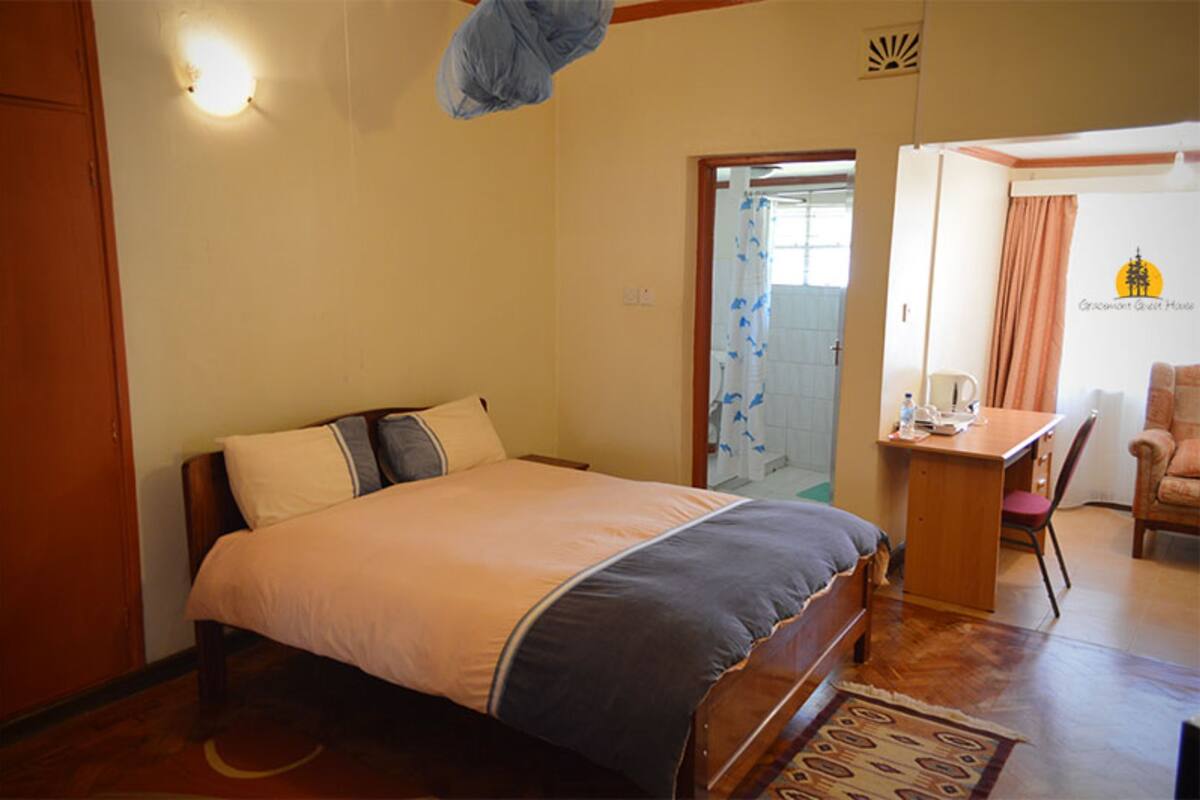 Quarto com cama, escrivaninha e cadeira. Há uma janela com cortinas e uma porta que dá para outro cômodo.