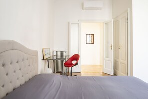 Un dormitorio con cabecera capitonada, cama con ropa de cama gris, una silla roja, un radiador y una puerta que da a otra habitación.