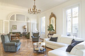 Een ruime woonkamer met een witte bank, gestreepte fauteuils, een ronde salontafel en een kroonluchter.