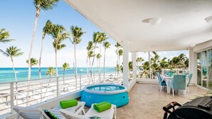 Balcón con piscina, sillas de descanso y zona de barbacoa con vistas a una playa con palmeras y el océano.