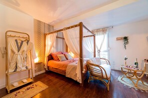 A four-poster bed with a canopy, a wicker chair, a small round table with a bottle and glasses, and a bedside table with a plant.