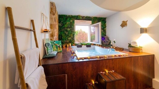 Ett spa-rum med ett badkar i trä, ett fönster med utsikt och en vägg dekorerad med grönska.