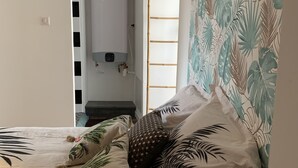 Une chambre à coucher avec un lit, des oreillers à motifs tropicaux, un radiateur et une fenêtre donnant sur un escalier.