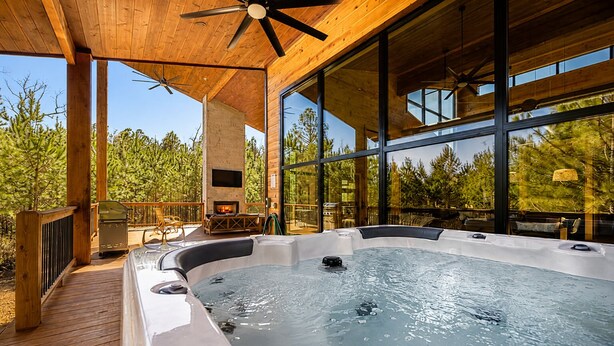 A hot tub on a wooden deck with a fireplace, large windows, and a view of trees. - Sunlit Spaces • 6-Bunk Room • Big Porch Fun (Broken Bow)