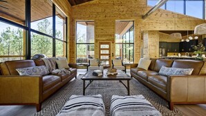 A spacious living room with leather sofas, a central coffee table, and ottomans. Large windows provide natural light and views of the outdoors. - Sunlit Spaces • 6-Bunk Room • Big Porch Fun (Broken Bow)