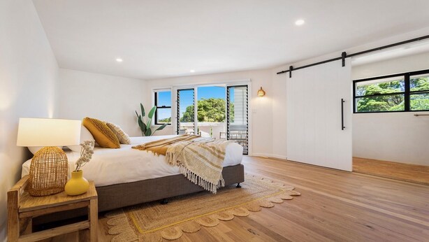 A modern bedroom with a large bed, a bedside table with a lamp, and a view of greenery through the windows. - Le Dog Beach Club - chic home with pool (Byron Bay)
