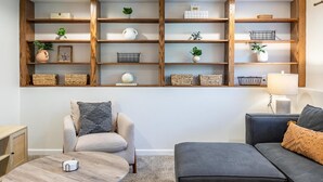 A living room with a sofa, armchair, and coffee table. There is a wooden shelf unit above the sofa with various decorative items. - Cozy, Clean and Bright 4BR / 2BA Home! Welcome! (Sterling Heights)