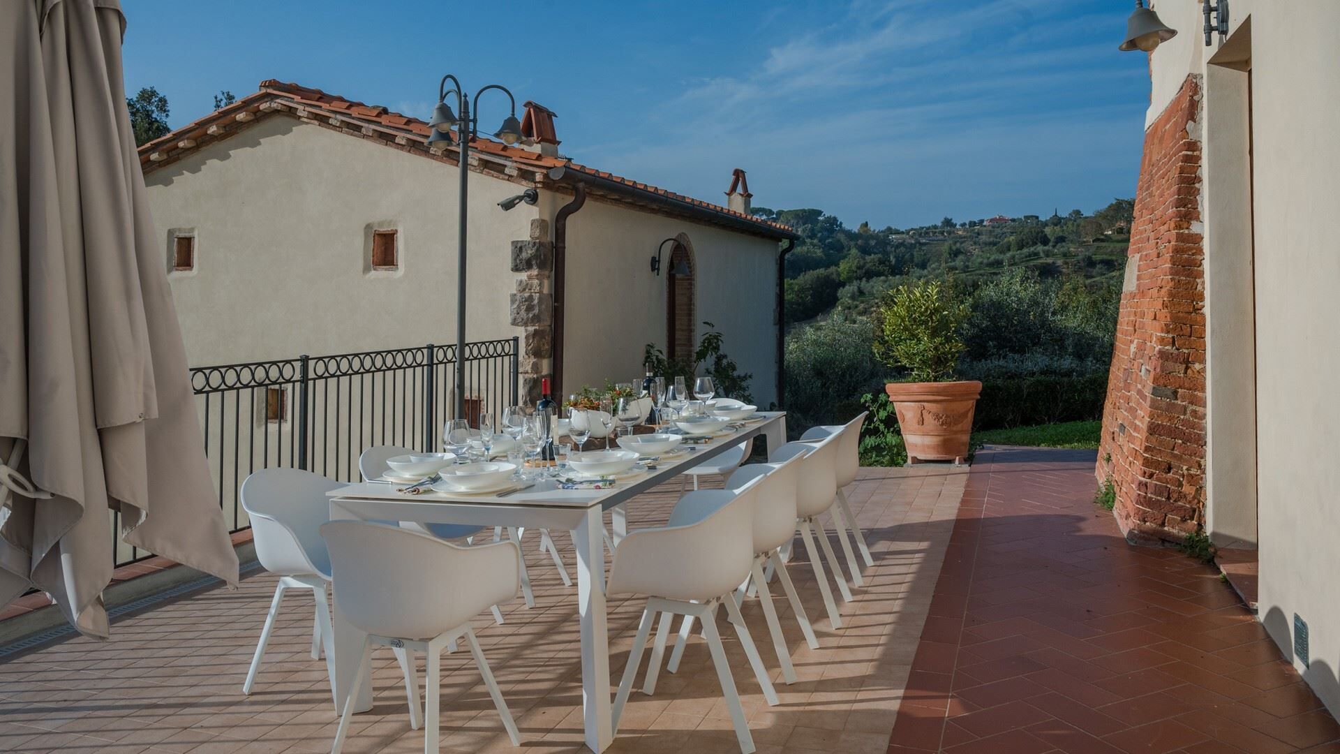Un espace de restauration en plein air avec une table dressée pour six personnes, entourée de chaises blanches, sur une terrasse offrant une vue sur une colline.