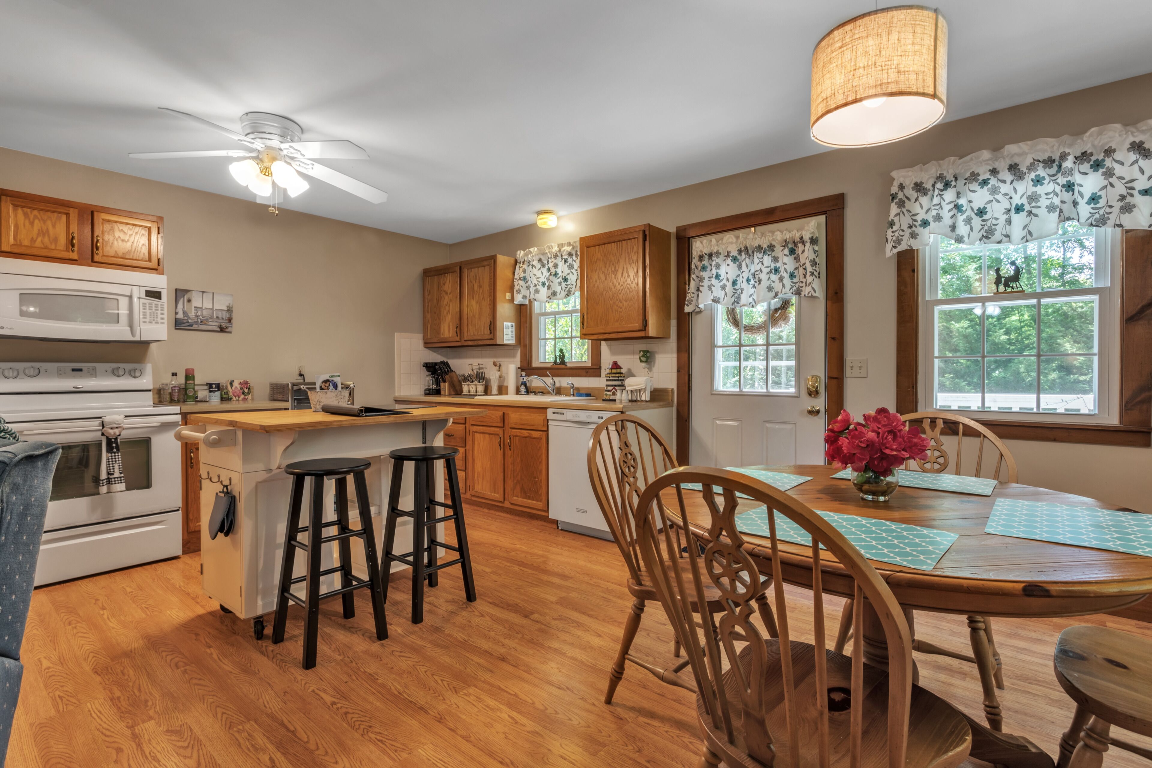 A kitchen with wooden cabinets, a dining table with chairs, a ceiling fan, and a window with a view of greenery.
