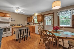 A kitchen with wooden cabinets, a dining table with chairs, a ceiling fan, and a window with a view of greenery.