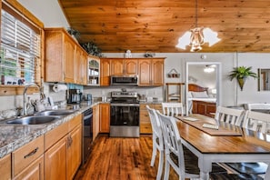 A kitchen with wooden cabinets, stainless steel appliances, and a dining area with white chairs. - Roundtop Mountain Ridge 2 Bedroom Cabin by RedAwning (Sevierville)