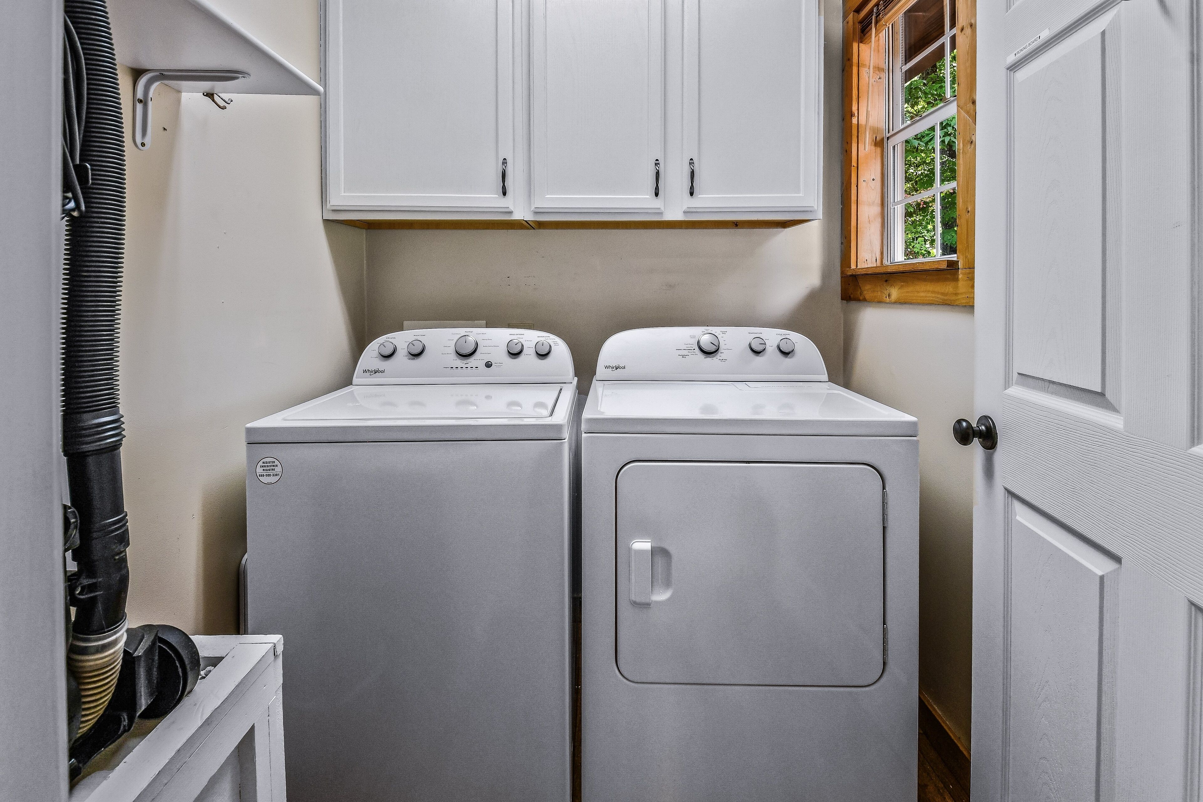 A laundry room with two white washers and dryers, white cabinets above, and a window with a view of greenery.