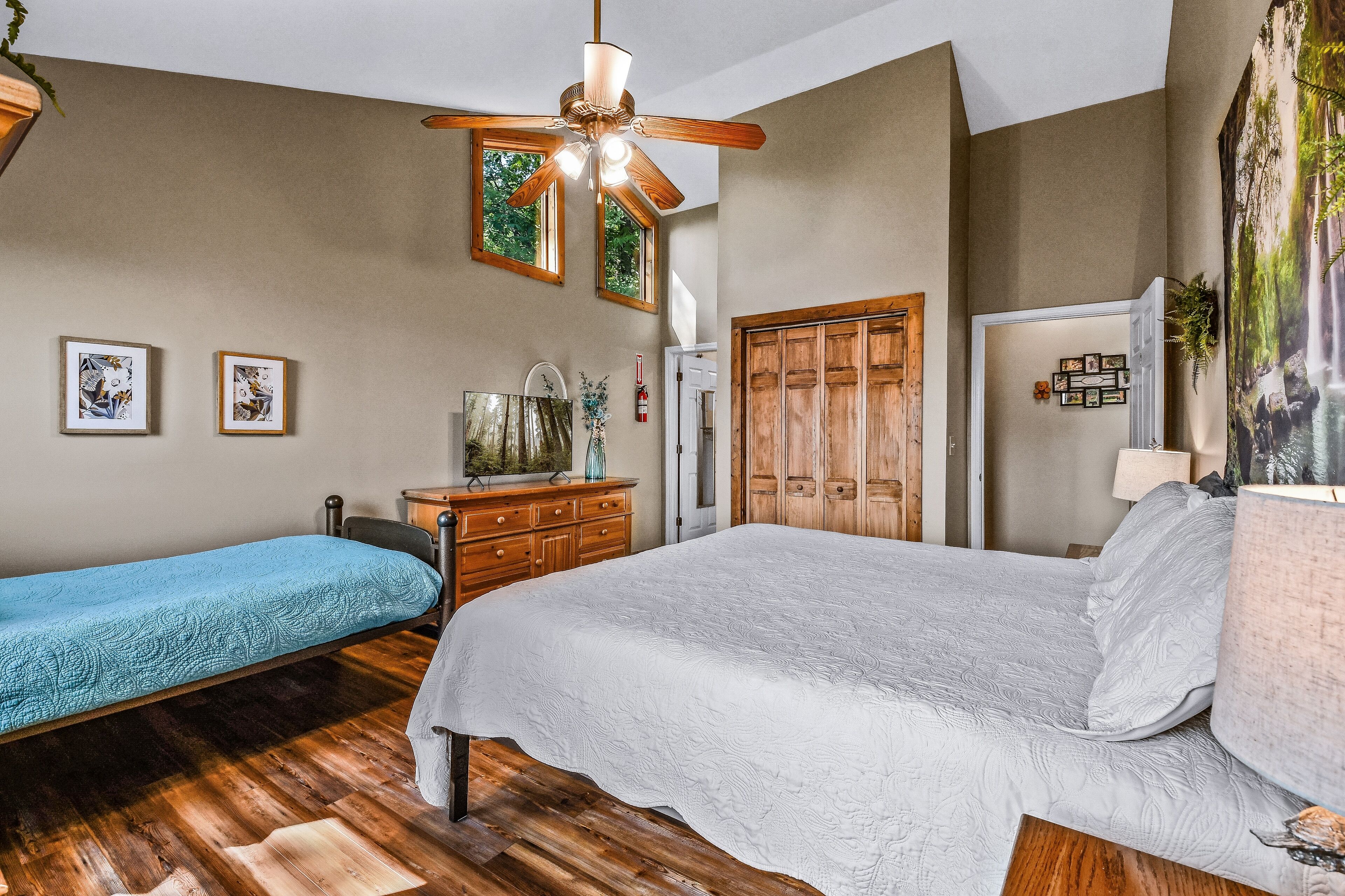 A bedroom with a large bed, a smaller bed, wooden flooring, a ceiling fan, and a wardrobe.