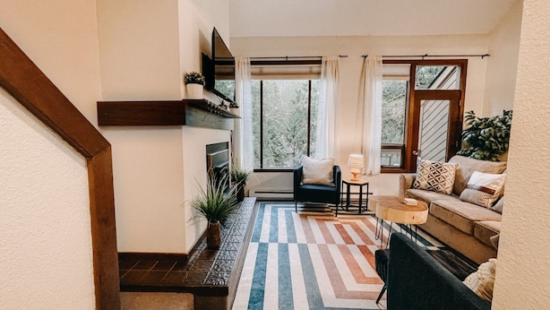 Un salon moderne avec un canapé, une table basse et une cheminée. Un escalier se trouve à gauche et une fenêtre à droite.