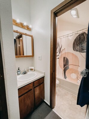 Un baño con un lavabo blanco, armarios de madera, un espejo con luces y una cortina de ducha con diseños abstractos.