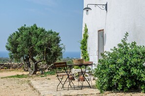 Un patio de piedra con mesa y sillas, un edificio blanco con una puerta y vistas al mar.