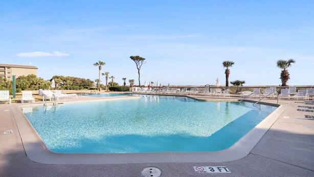 Ein rechteckiger Swimmingpool mit klarem, blauem Wasser, umgeben von weißen Liegestühlen und Palmen.