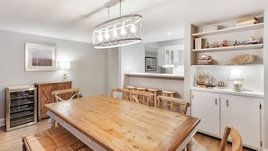 A dining area with a wooden table and chairs, a chandelier, a white cabinet with decorative items, and a kitchen area in the background.