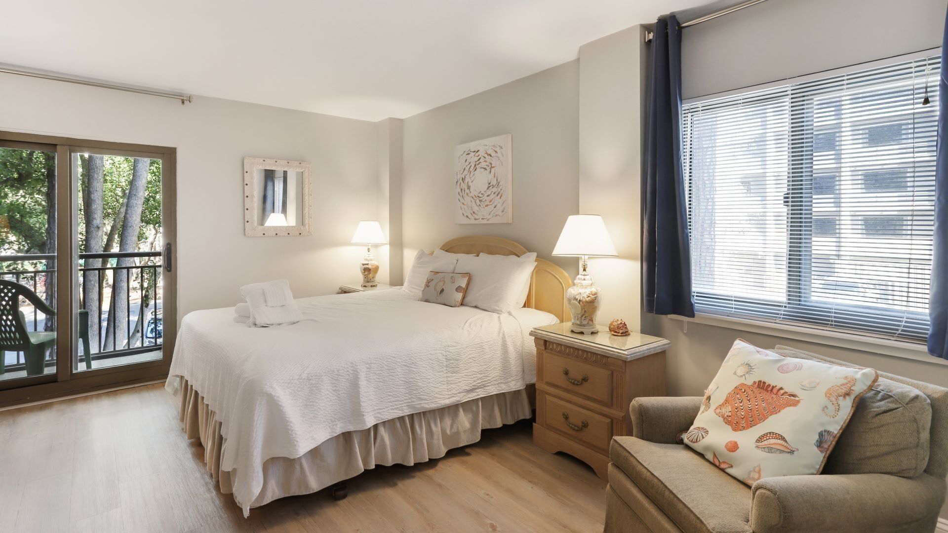 A bedroom with a bed, bedside tables, a chair, a window with blinds, and a balcony view.