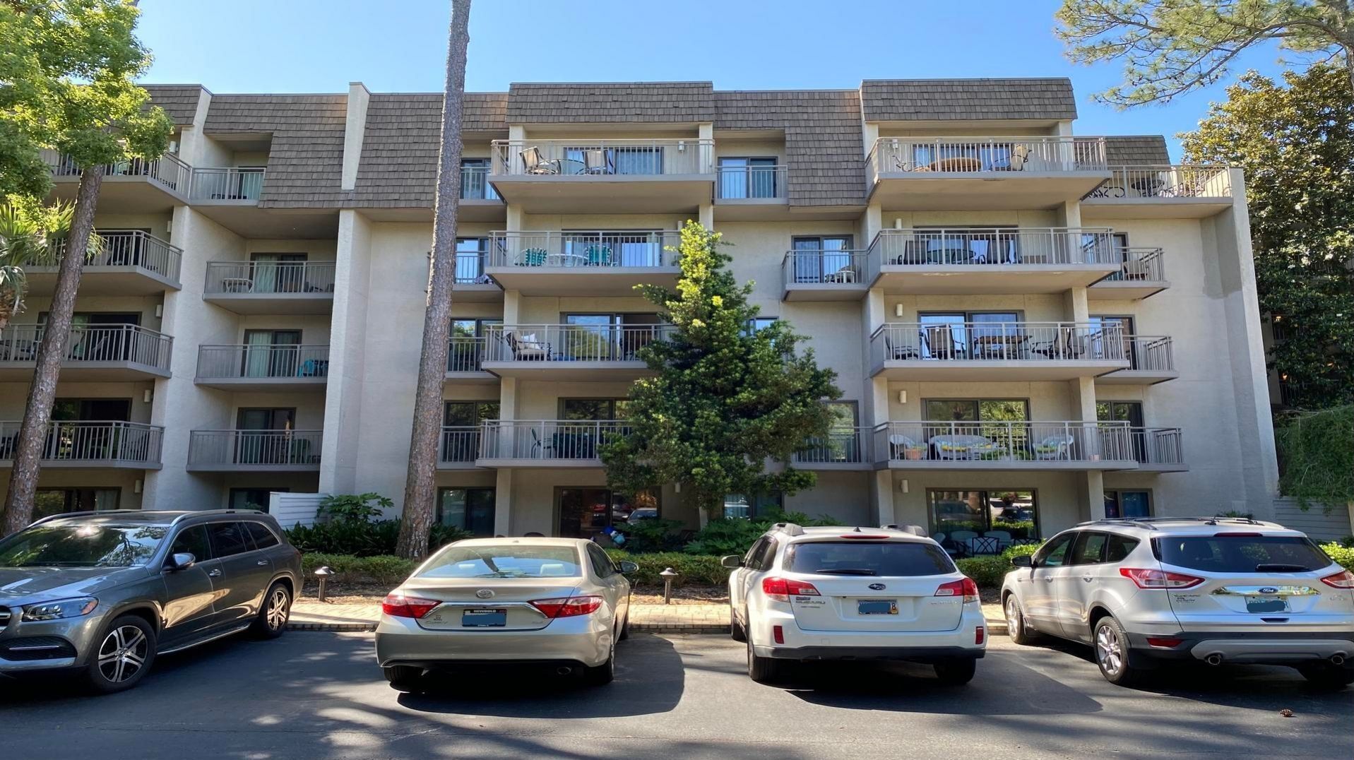 A multi-story apartment building with balconies, parking spaces, and a tree in front.