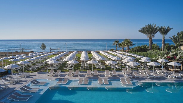 Ein Poolbereich mit Liegestühlen und Sonnenschirmen, ein strahlend blauer Himmel und das Meer im Hintergrund.