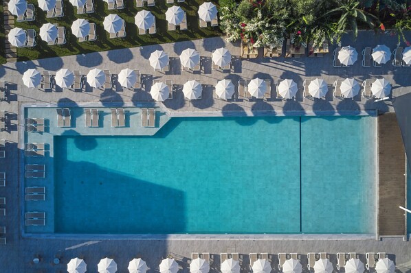 An aerial view of a swimming pool surrounded by sun loungers and umbrellas, with a clear blue pool and a landscaped area with trees and flowers.