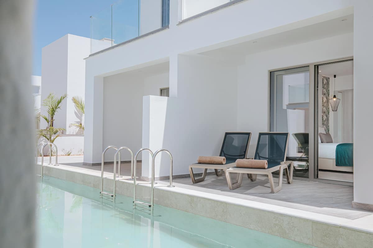 Un área de piscina moderna con agua azul cristalina, dos sillas de descanso con toallas y una puerta de cristal que da a una habitación.