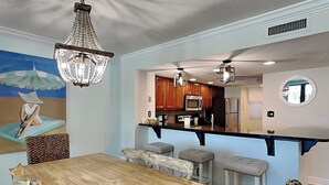 A dining area with a wooden table and chairs, a chandelier, and a kitchen in the background.