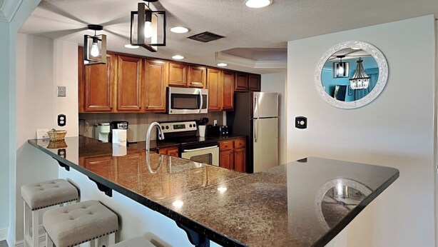 A modern kitchen with wooden cabinets, a granite countertop, a built-in microwave, a stainless steel refrigerator, and a round mirror on the wall.