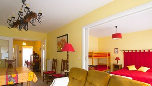 Une pièce comprenant un canapé, une table recouverte d’une nappe à motifs floraux, une lampe rouge et un lit superposé avec une literie rouge.