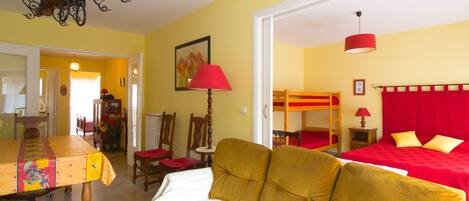 Una habitación con sofá, una mesa con mantel floral, una lámpara roja y una cama litera con ropa de cama roja.