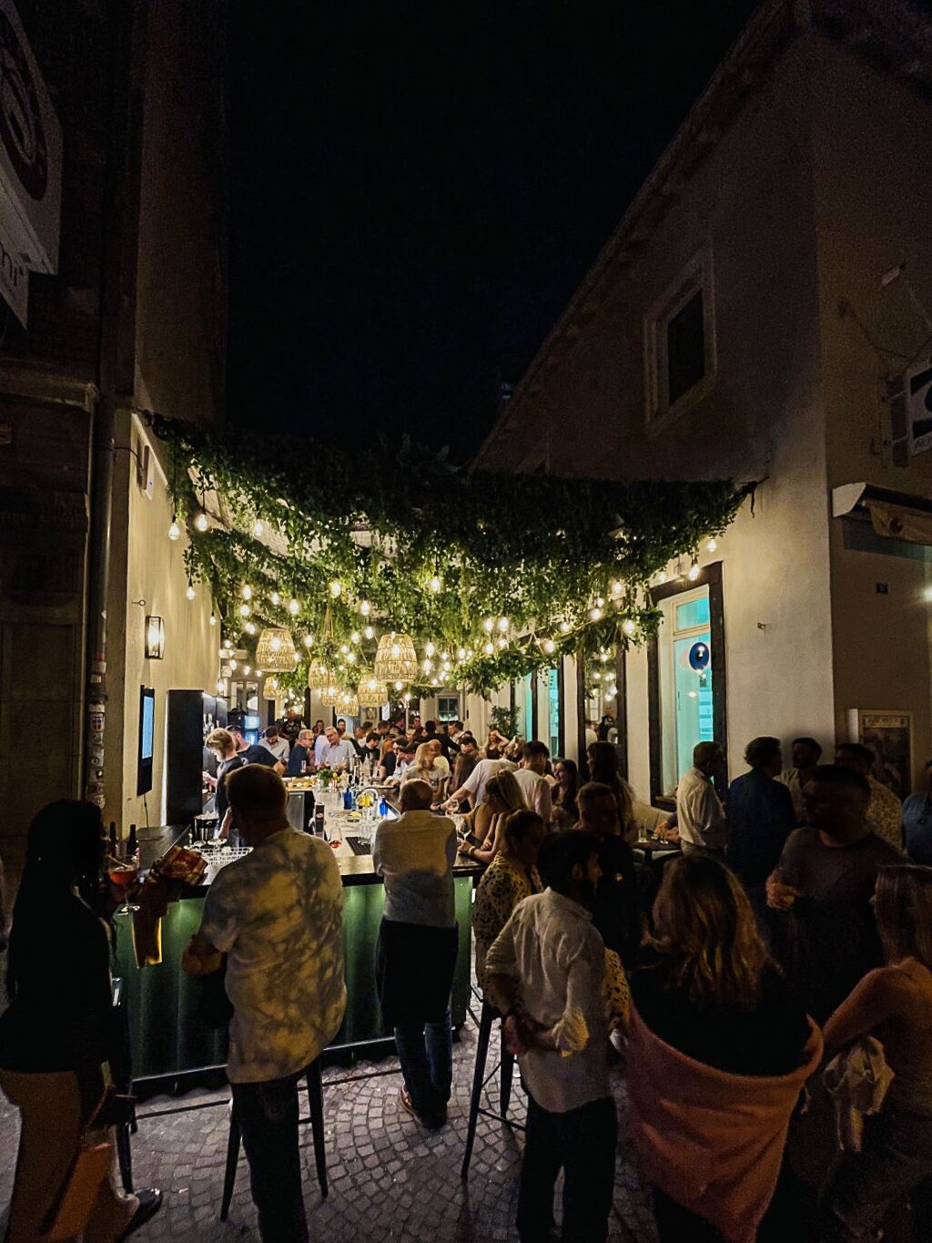 Uma área de bar ao ar livre com gente, balcão e um cenário de vegetação e luzinhas.