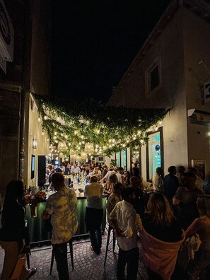 An outdoor bar area with a crowd of people, a bar counter, and a backdrop of greenery and string lights. - Hotel Fuchs (Saarbrücken)