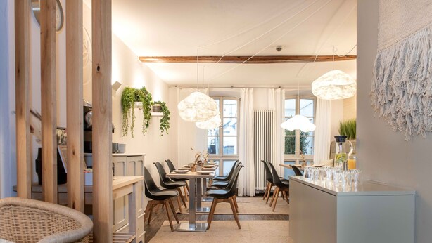 A modern dining area with a long table, chairs, hanging plants, and pendant lights. - Hotel Fuchs (Saarbrücken)