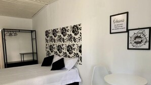 Un dormitorio con una cama, una mesita pequeña y dos sillas. Se observa una pared con diseño floral y dos cuadros enmarcados.