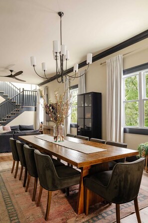 Un comedor con una mesa de madera, sillas negras, una araña y vistas a una escalera y follaje exterior.