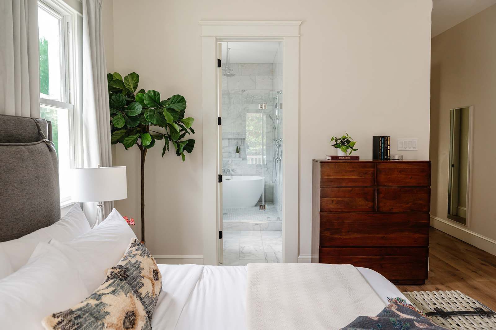 Une chambre à coucher comprenant un lit, une commode, une plante et donnant sur une salle de bain avec une douche et une baignoire.