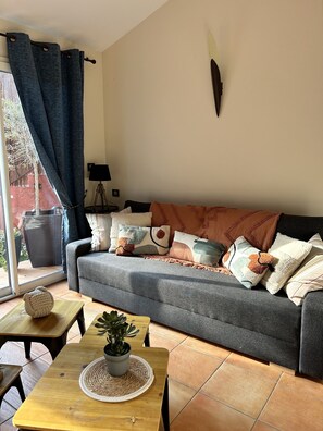 A cozy living room with a grey sofa, a wooden coffee table with a plant, and a view through a window with curtains.