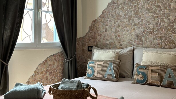 A bedroom with a stone headboard, a bed with pillows, a window with curtains, and a basket on the floor. - Grenache (Collioure)