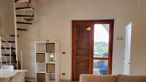 A living room with a sofa, a dining table with chairs, a white shelving unit, a hanging light fixture, and a view through a door. - Vacation home with panoramic views by the sea (San Nicola Arcella)
