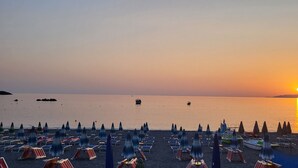 Een strand met rijen parasols en ligbedden, boten op het water en een zonsondergang op de achtergrond.