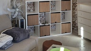 Een woonkamer met een metselwerk wand, een flatscreen televisie, een wit wandrek met rieten manden, een grijze bank en een witte salontafel met een groene schaal erop.