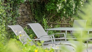 An outdoor seating area with metal chairs and a table, surrounded by greenery and a stone wall.