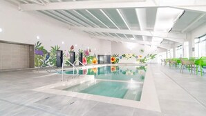 Una piscina interior con suelo de baldosas, rodeada de sillas y mesas verdes, y paredes decoradas con murales florales.