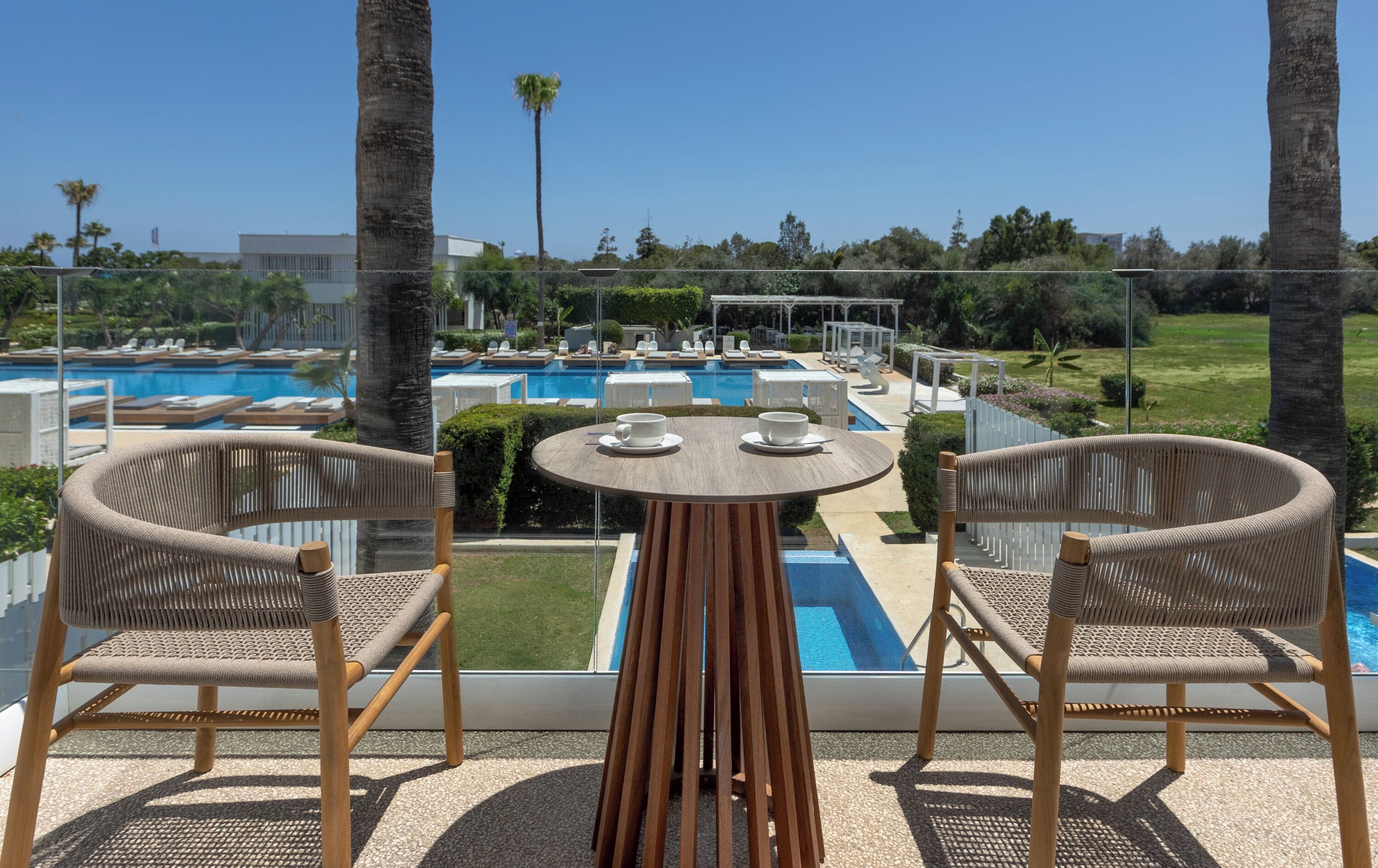Un balcón con sillas de mimbre y una mesa con vistas a una zona de piscina que cuenta con varias piscinas y tumbonas.