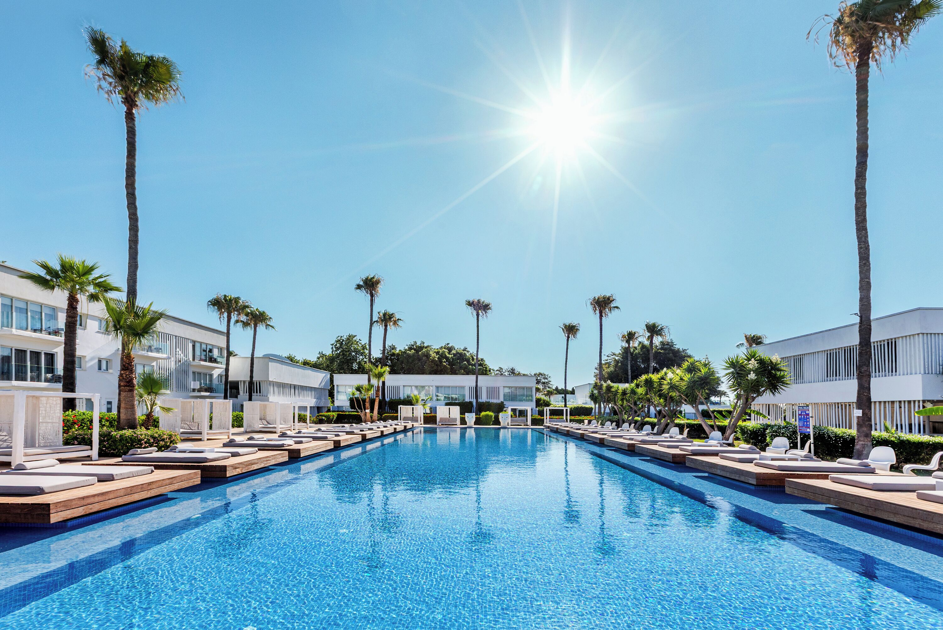 Piscina com espreguiçadeiras e palmeiras, cercada por edifícios modernos.