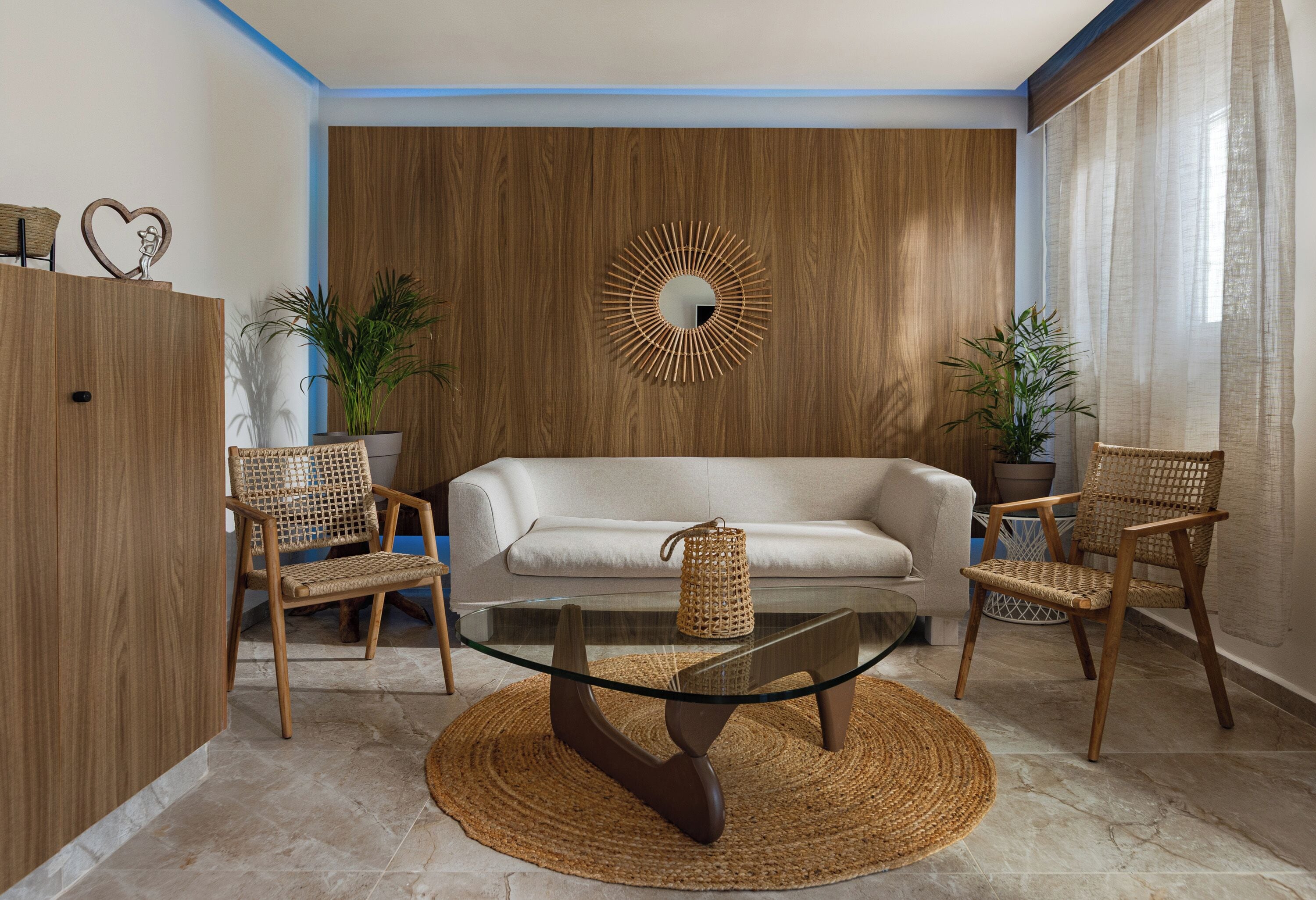 Ein modernes Wohnzimmer mit einem Glastisch, Korbstühlen, einem weißen Sofa und einer Wand mit Holzvertäfelung und einem runden Spiegel.