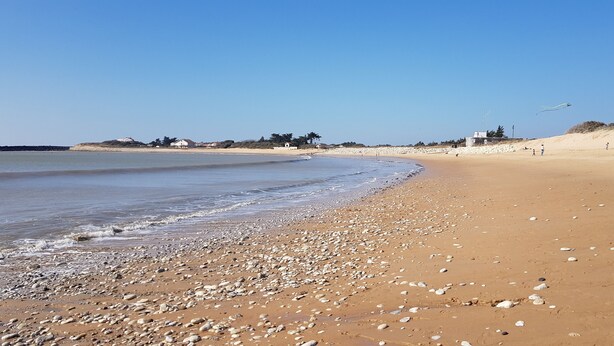 Ein Strand mit Kieselsteinen und Sand, ein klarer Himmel und einige Gebäude in der Ferne.